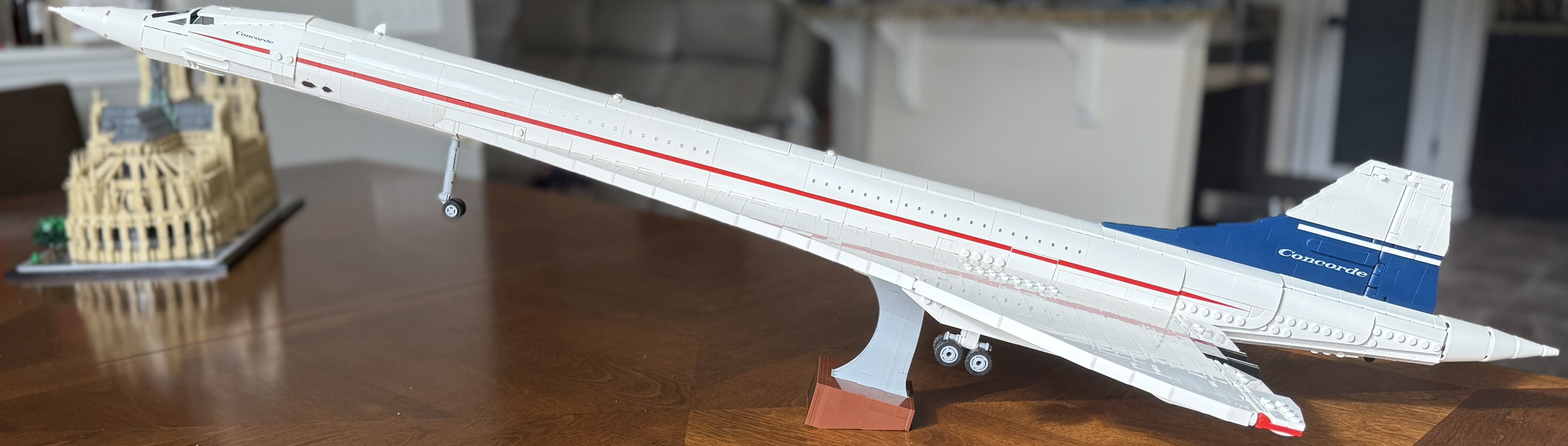 A detailed LEGO<sup>®</sup> model of the Concorde supersonic airliner is displayed on a wooden table. The model is predominantly white with a red stripe running along its fuselage and a blue tail fin featuring the word 'Concorde.' The nose is in a downward position, and the landing gear is extended. In the background, there is another LEGO<sup>®</sup> model of Notre-Dame Cathedral in Paris, placed on a black base.