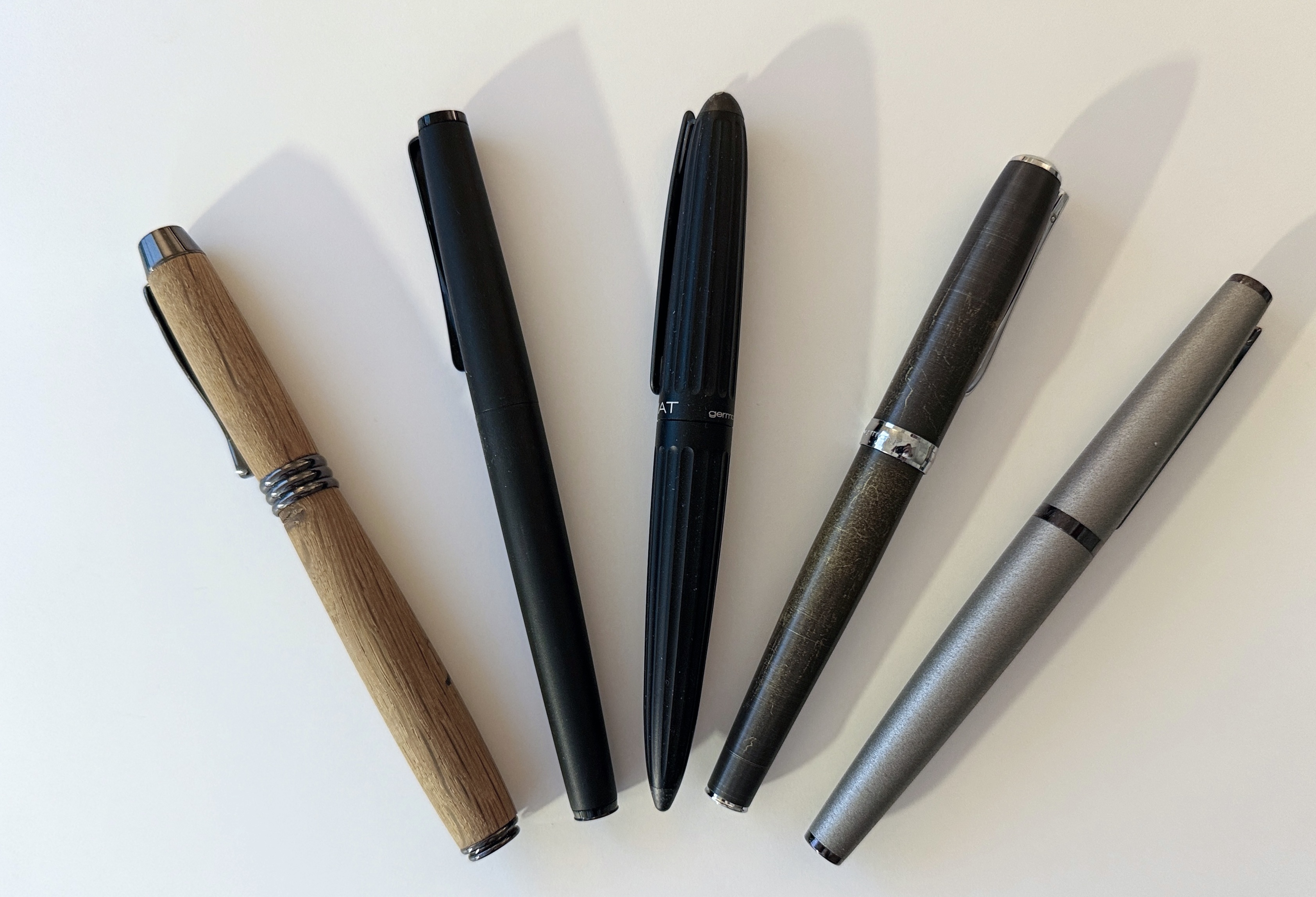 This image shows five pens arranged in a row on a white surface. Each pen has a distinct design and material, including a wooden pen on the left, a matte black pen, a ribbed black pen in the center, a metallic pen with a textured surface, and a sleek silver pen on the right.