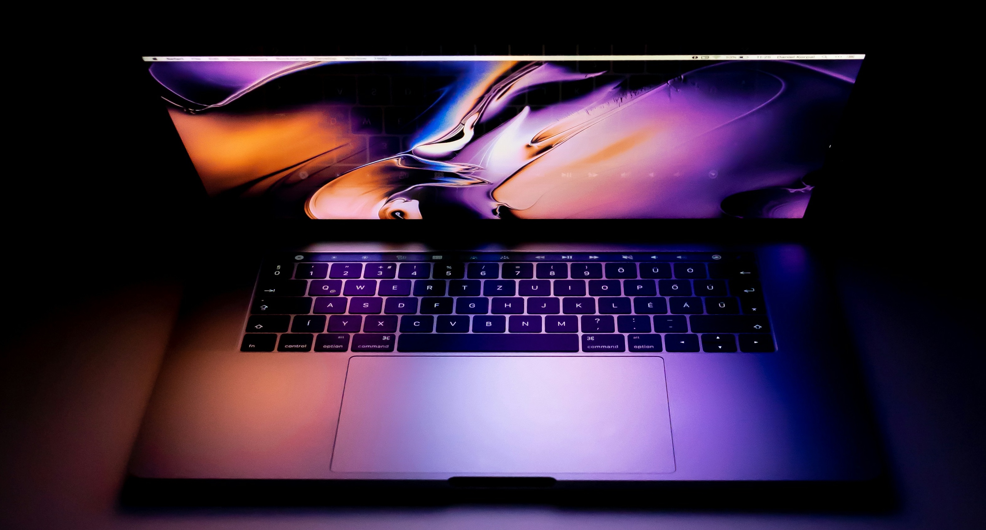 This is an artistic photograph of a MacBook laptop with a vibrant, abstract wallpaper displayed on the screen. The lighting creates a dramatic effect, with a mix of purple, orange, and blue hues reflecting on the keyboard and trackpad. The dark surroundings emphasize the illuminated laptop, making it the focal point of the image.