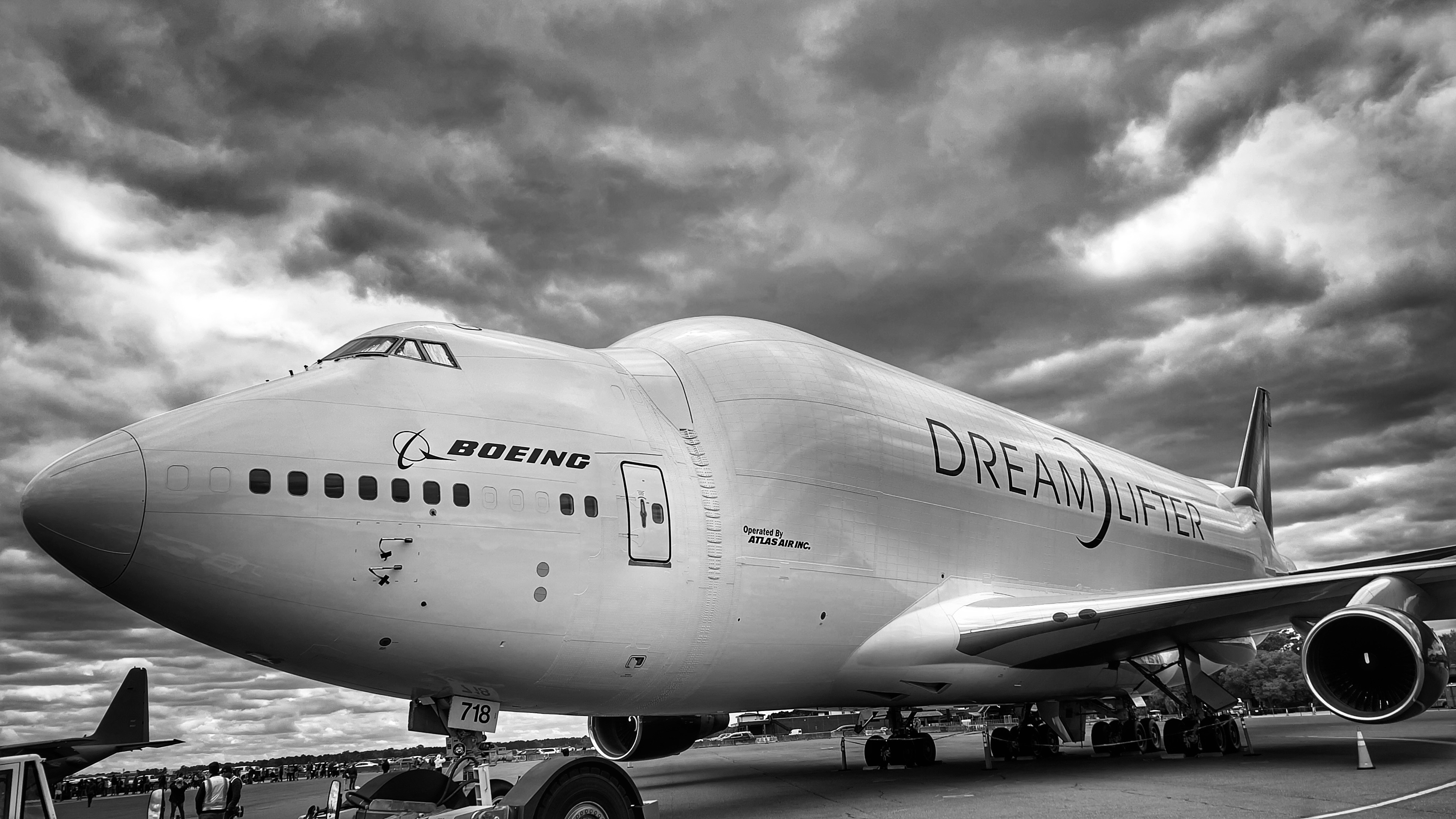 Black and white image of one of Boeing's Dreamlifters, a modified 747 aircraft used for transporting parts for Boeing's 787 assembly facilities in Charleston, South Carolina and Seattle, Washington. The white plane is set against dark storm clouds.